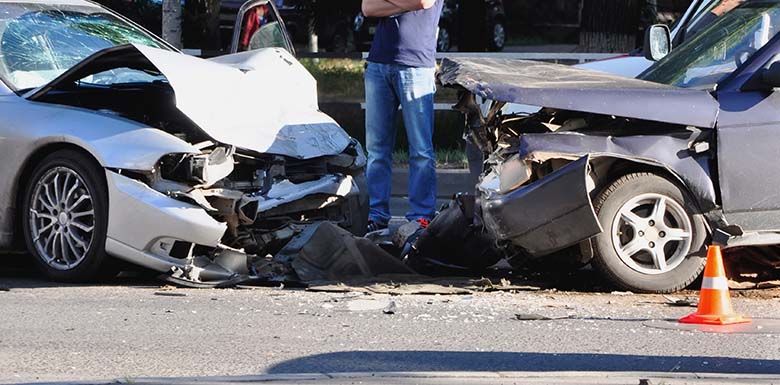 Two cars with significant front-end damage after a head-on collision. A person stands nearby, facing away. An orange traffic cone is in front of the black car. Debris is scattered on the road.