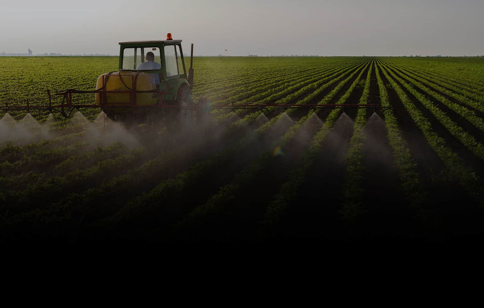 A tractor sprays crops with pesticides in a vast, evenly plowed field under a clear sky.