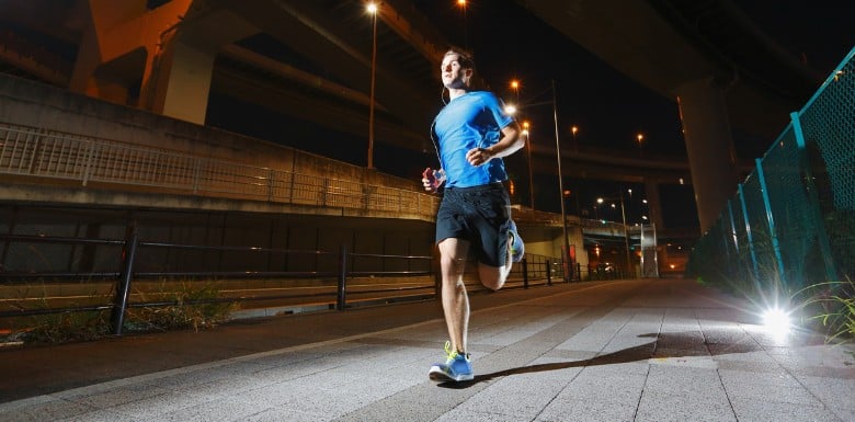 A man wearing a blue shirt and black shorts runs on a paved path at night under an overpass with streetlights illuminating the scene.