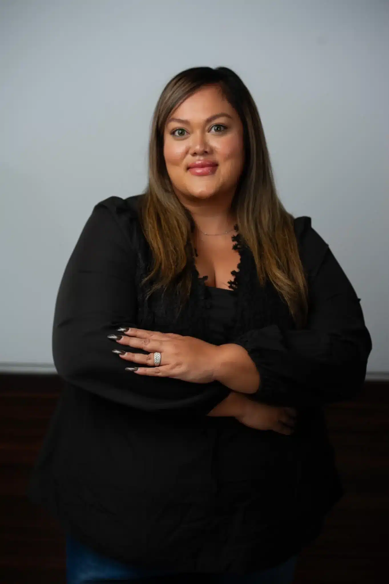 A woman with long brown hair wearing a black blouse stands with arms crossed, smiling at the camera against a plain background.