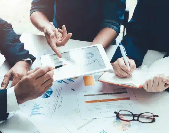 Three people review charts and graphs on a tablet and paper documents at a table, discussing data and taking notes. Glasses rest on the table beside them.