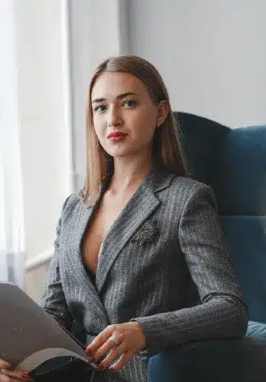 A woman with straight brown hair, wearing a gray pinstripe blazer, sits on a blue chair holding a magazine in her lap.
