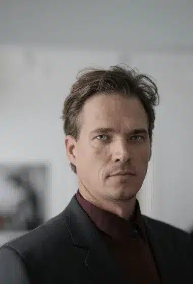A man with short brown hair and blue eyes wearing a dark suit and maroon shirt stands indoors, looking directly at the camera.