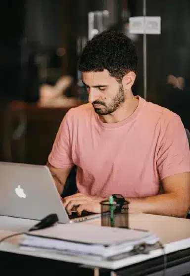 A man wearing a pink T-shirt is sitting at a desk working on a laptop. There are papers and office supplies on the desk in front of him.